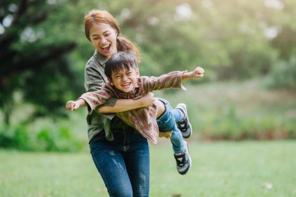 A mom holds her son off the ground, pretending he's an airplane. They are playing outside, surrounded by trees.