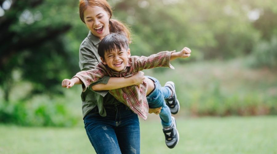 A mom holds her son off the ground, pretending he's an airplane. They are playing outside, surrounded by trees.