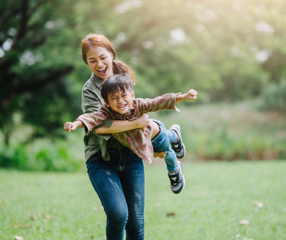 A mom holds her son off the ground, pretending he's an airplane. They are playing outside, surrounded by trees.