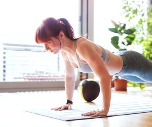 A woman wearing athletic clothes holds a plank position on a mat. A weighted ball and potted plants are in the background.