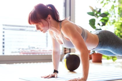 A woman wearing athletic clothes holds a plank position on a mat. A weighted ball and potted plants are in the background.