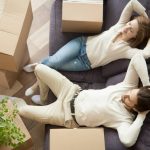 A couple sitting on a purple couch, lounging with their hands behind their heads and surrounded by a handful of cardboard boxes.