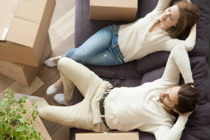 A couple sitting on a purple couch, lounging with their hands behind their heads and surrounded by a handful of cardboard boxes.