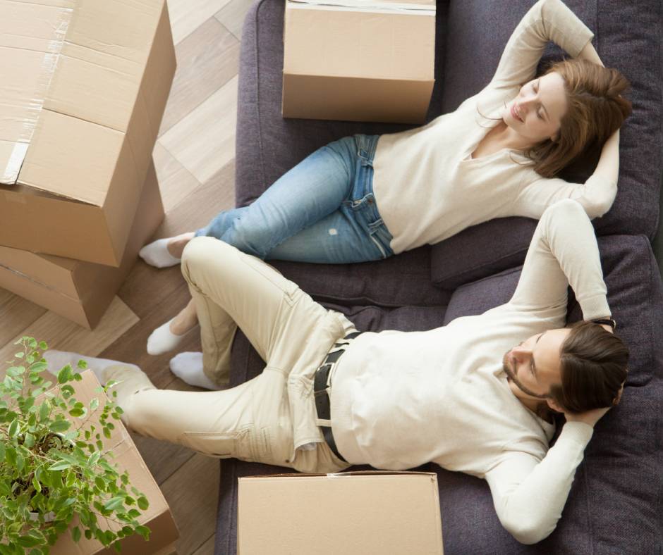 A couple sitting on a purple couch, lounging with their hands behind their heads and surrounded by a handful of cardboard boxes.