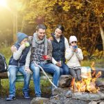 A four-person family sits on a log in a line next to a fire pit, surrounded by a forest of fall colors.