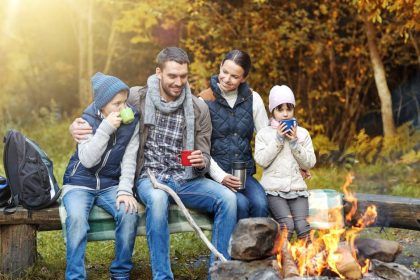 A four-person family sits on a log in a line next to a fire pit, surrounded by a forest of fall colors.