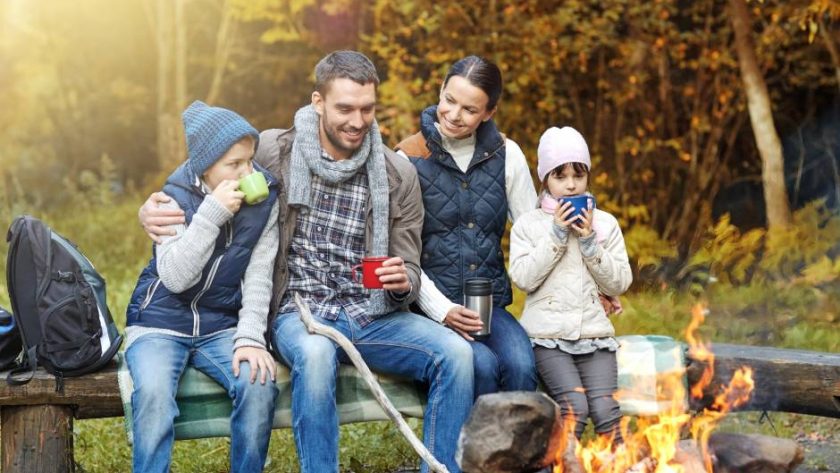 A four-person family sits on a log in a line next to a fire pit, surrounded by a forest of fall colors.