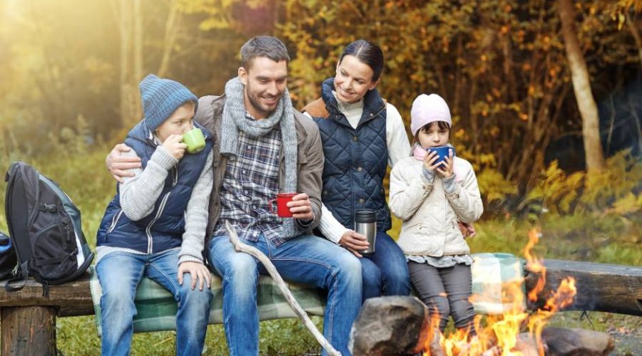 A four-person family sits on a log in a line next to a fire pit, surrounded by a forest of fall colors.