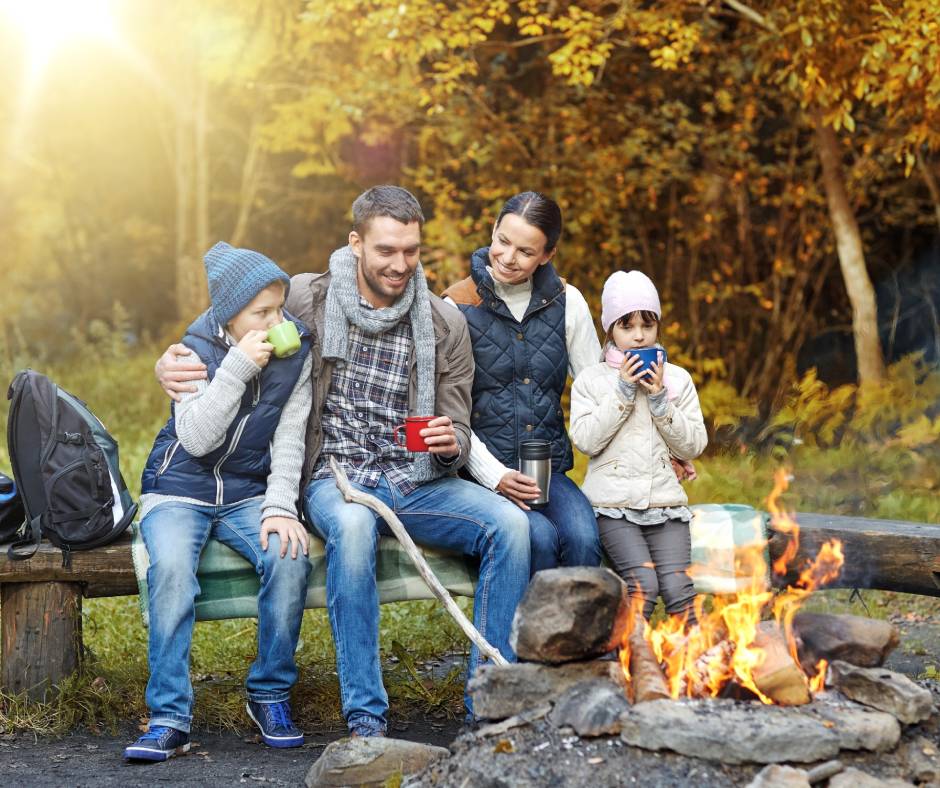 A four-person family sits on a log in a line next to a fire pit, surrounded by a forest of fall colors.