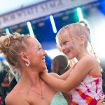 A mom carrying her daughter at a music festival. They both have glitter face paint and colorful hair accessories.