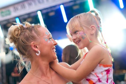 A mom carrying her daughter at a music festival. They both have glitter face paint and colorful hair accessories.