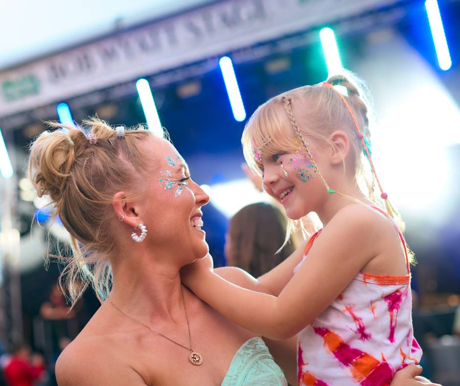 A mom carrying her daughter at a music festival. They both have glitter face paint and colorful hair accessories.