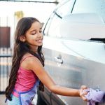 A young girl with long brown hair smiling while using a purple cloth to wash the outside of a vehicle.