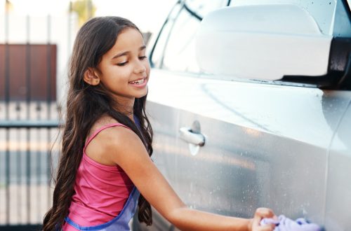 A young girl with long brown hair smiling while using a purple cloth to wash the outside of a vehicle.