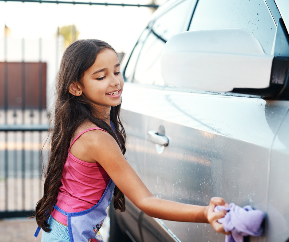 A young girl with long brown hair smiling while using a purple cloth to wash the outside of a vehicle.