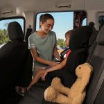 A young mother buckling up her child in the backseat of a car with a brown rabbit stuffed animal next to him.