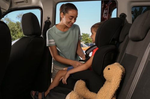 A young mother buckling up her child in the backseat of a car with a brown rabbit stuffed animal next to him.