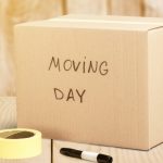 A taped-shut cardboard box reads "moving day" in black marker. It sits next to a black marker and a roll of packing tape.