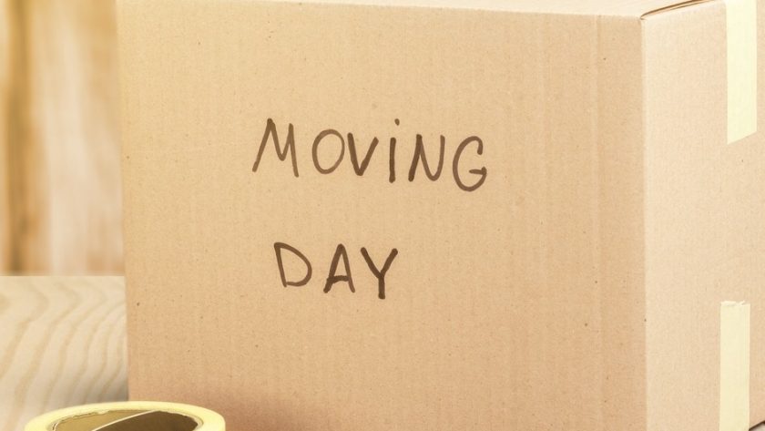 A taped-shut cardboard box reads "moving day" in black marker. It sits next to a black marker and a roll of packing tape.