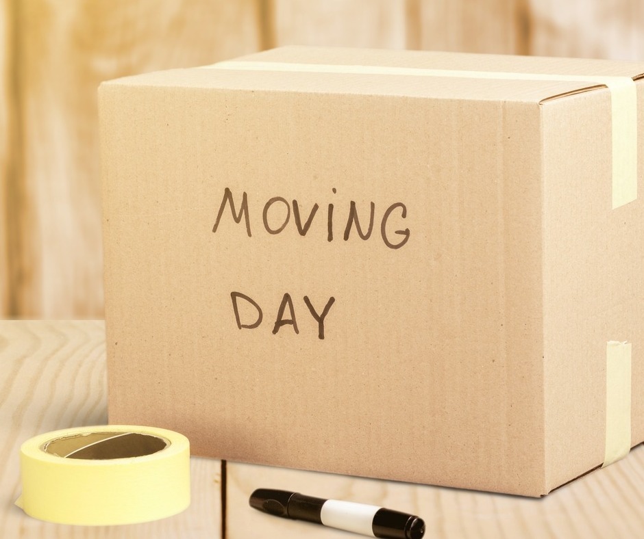 A taped-shut cardboard box reads "moving day" in black marker. It sits next to a black marker and a roll of packing tape.