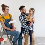 Two happy young parents with their daughter holding a paintbrush. The mother sits on a step ladder and holds a paint roller.