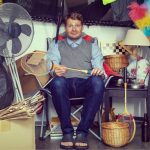 A man sitting down with a tennis racket in his hands looking emotional. He's surrounded by clutter.