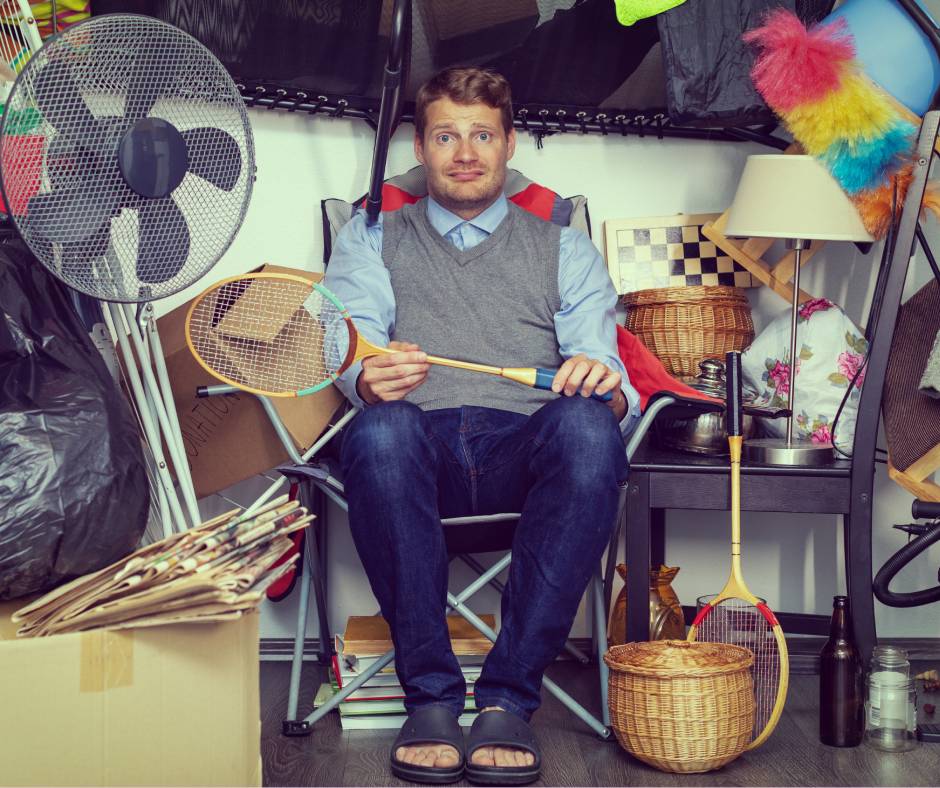 A man sitting down with a tennis racket in his hands looking emotional. He's surrounded by clutter.