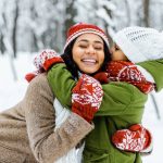 A young mother closely hugs her daughter in a snowy and open forest. She smiles as her daughter embraces her back.