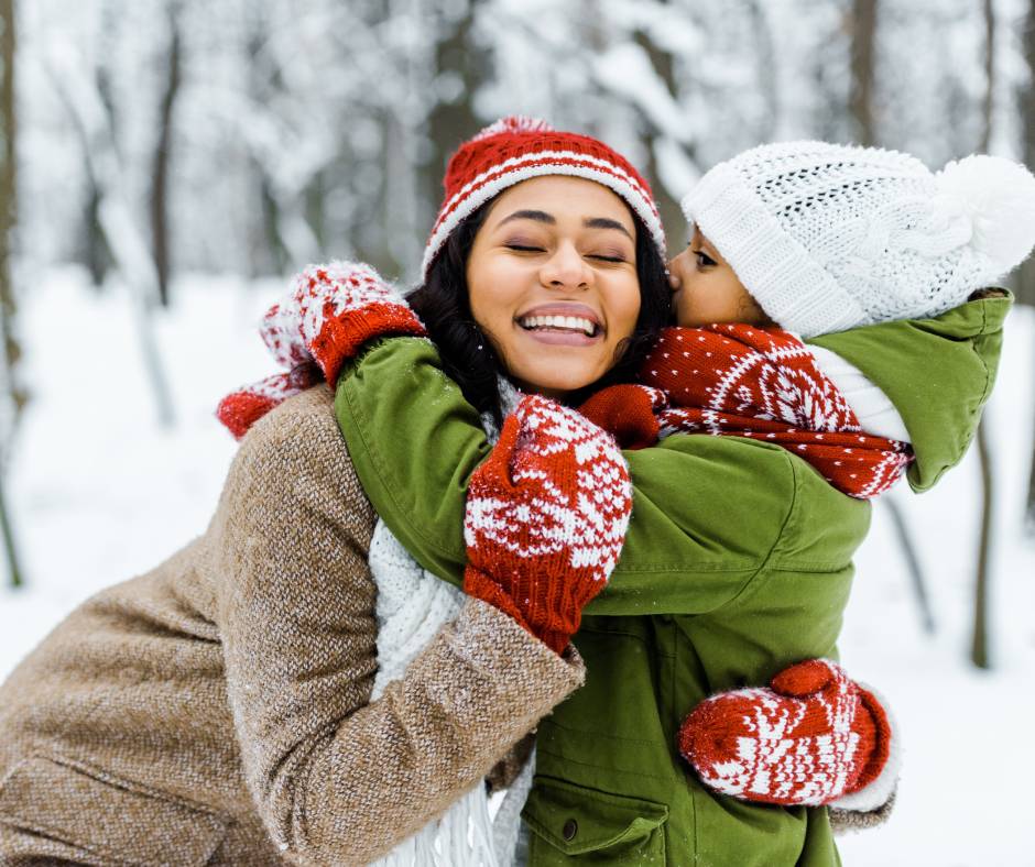 A young mother closely hugs her daughter in a snowy and open forest. She smiles as her daughter embraces her back.