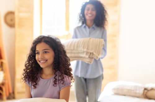 A young mother and her daughter carry clean blankets and smile in a brightly-lit bedroom.