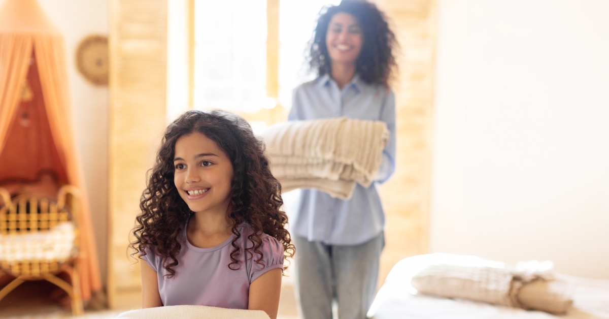 A young mother and her daughter carry clean blankets and smile in a brightly-lit bedroom.