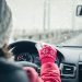 A woman in a winter hat and gloves driving her car. It is cloudy outside of the car as snow sticks to her windshield.