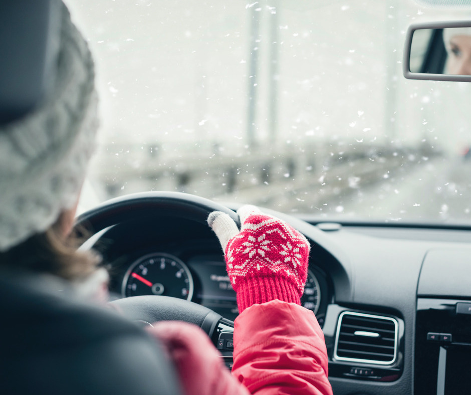 A woman in a winter hat and gloves driving her car. It is cloudy outside of the car as snow sticks to her windshield.