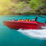 A red jet boat full of people wearing yellow life vests and blue helmets drives through water.