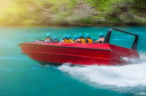 A red jet boat full of people wearing yellow life vests and blue helmets drives through water.