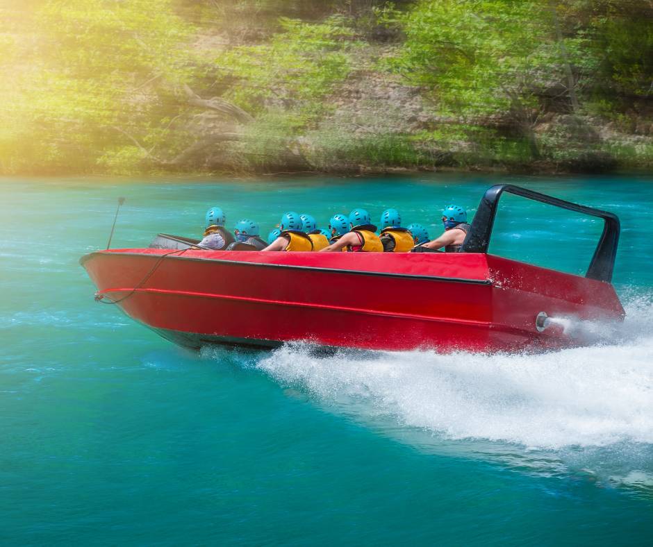 A red jet boat full of people wearing yellow life vests and blue helmets drives through water.