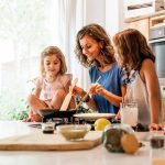 A middle-aged mother cooking in the kitchen with young daughters. She holds utensils in a pan on the stove.