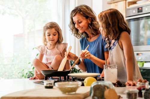 A middle-aged mother cooking in the kitchen with young daughters. She holds utensils in a pan on the stove.