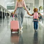 A mother and her child walking the halls of an airport. They're holding hands and the mom is rolling the luggage.