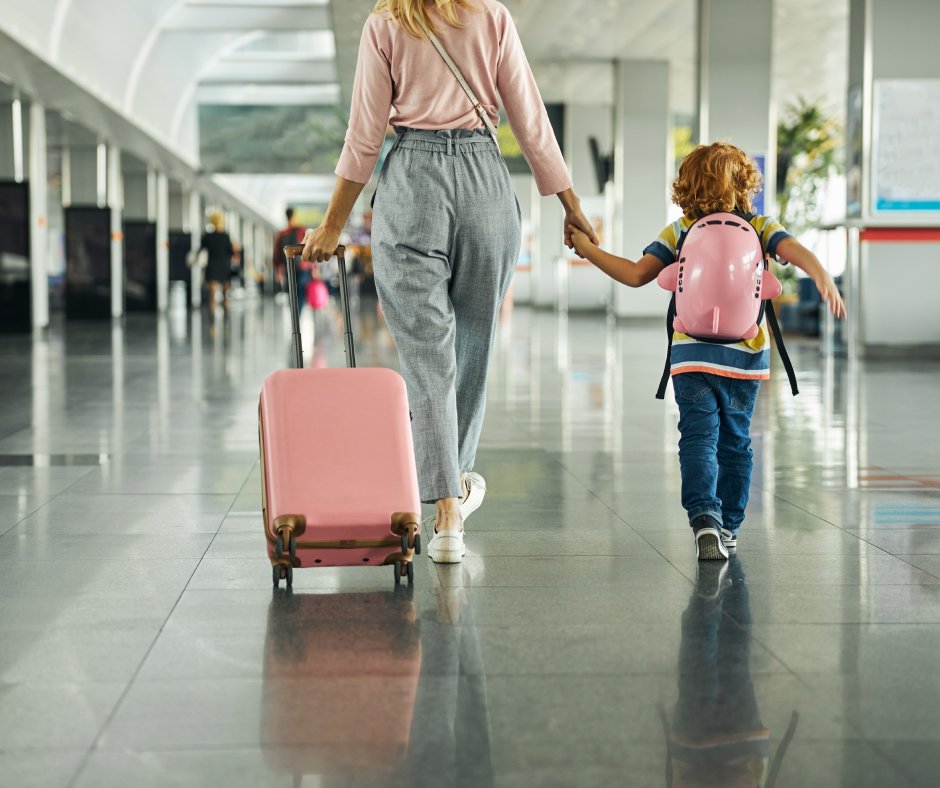 A mother and her child walking the halls of an airport. They're holding hands and the mom is rolling the luggage.