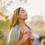 A woman has her eyes closed and both hands over her heart as she stands outside with her head slightly tilted upward.