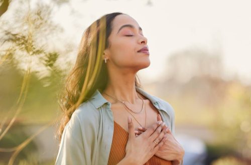 A woman has her eyes closed and both hands over her heart as she stands outside with her head slightly tilted upward.