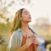 A woman has her eyes closed and both hands over her heart as she stands outside with her head slightly tilted upward.