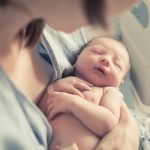 A close-up view shows a newborn being cradled in its mother's arms in a hospital bed. The mother wears a hospital gown.