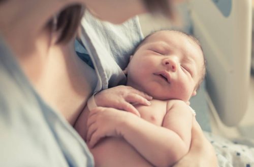 A close-up view shows a newborn being cradled in its mother's arms in a hospital bed. The mother wears a hospital gown.