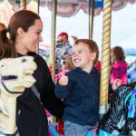 A mom and her son riding on a carousel. She is holding him by the back as he rides on a carousel horse.