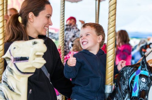 A mom and her son riding on a carousel. She is holding him by the back as he rides on a carousel horse.