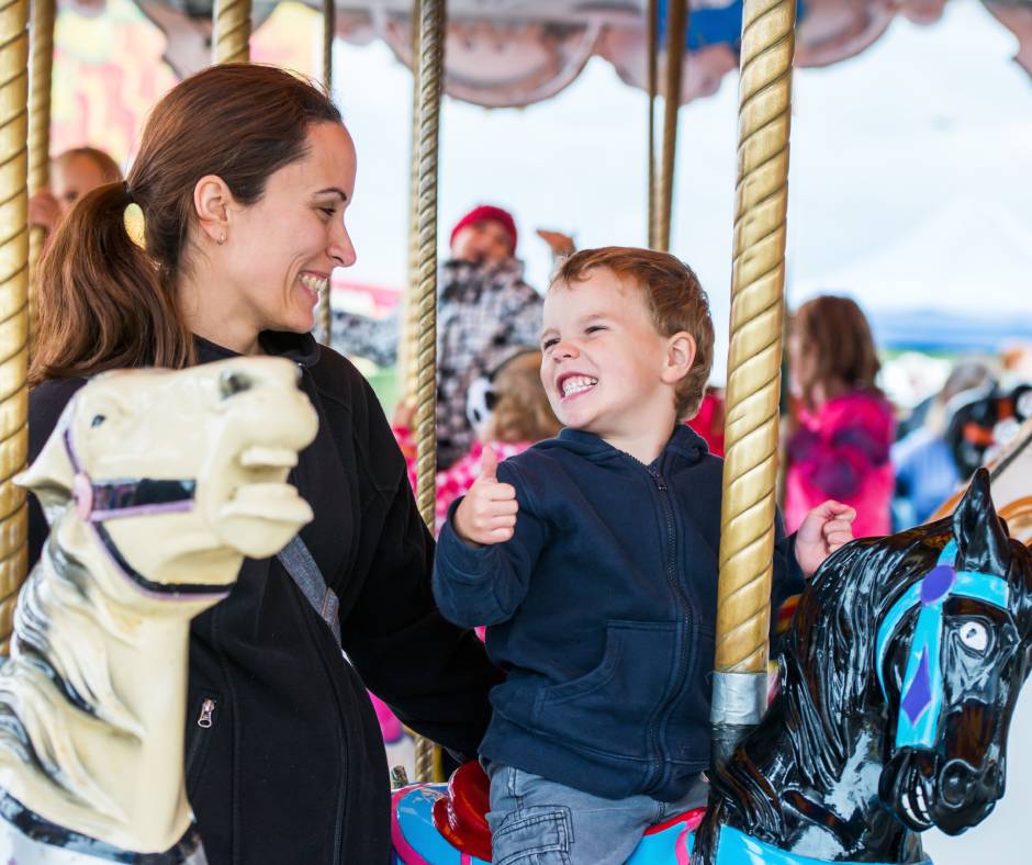 A mom and her son riding on a carousel. She is holding him by the back as he rides on a carousel horse.