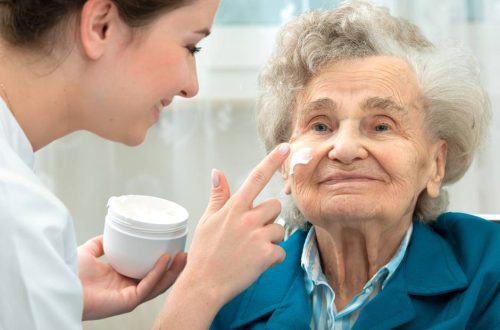 A woman putting facial cream on an elderly woman. The elderly woman is wearing a blue jacket.