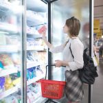 A woman opening a freezer door at the grocery store and selecting a frozen food item. She's holding a basket.